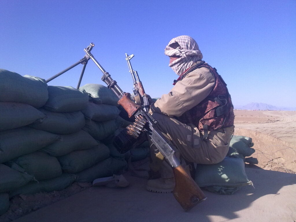 A vigilant security professional in a desert environment, wearing a shemagh and sitting behind sandbags with a mounted machine gun and a rifle