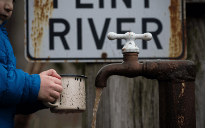 Child drinking from rusty tap with Flint River sign, highlighting Flint water crisis and lead poisoning.